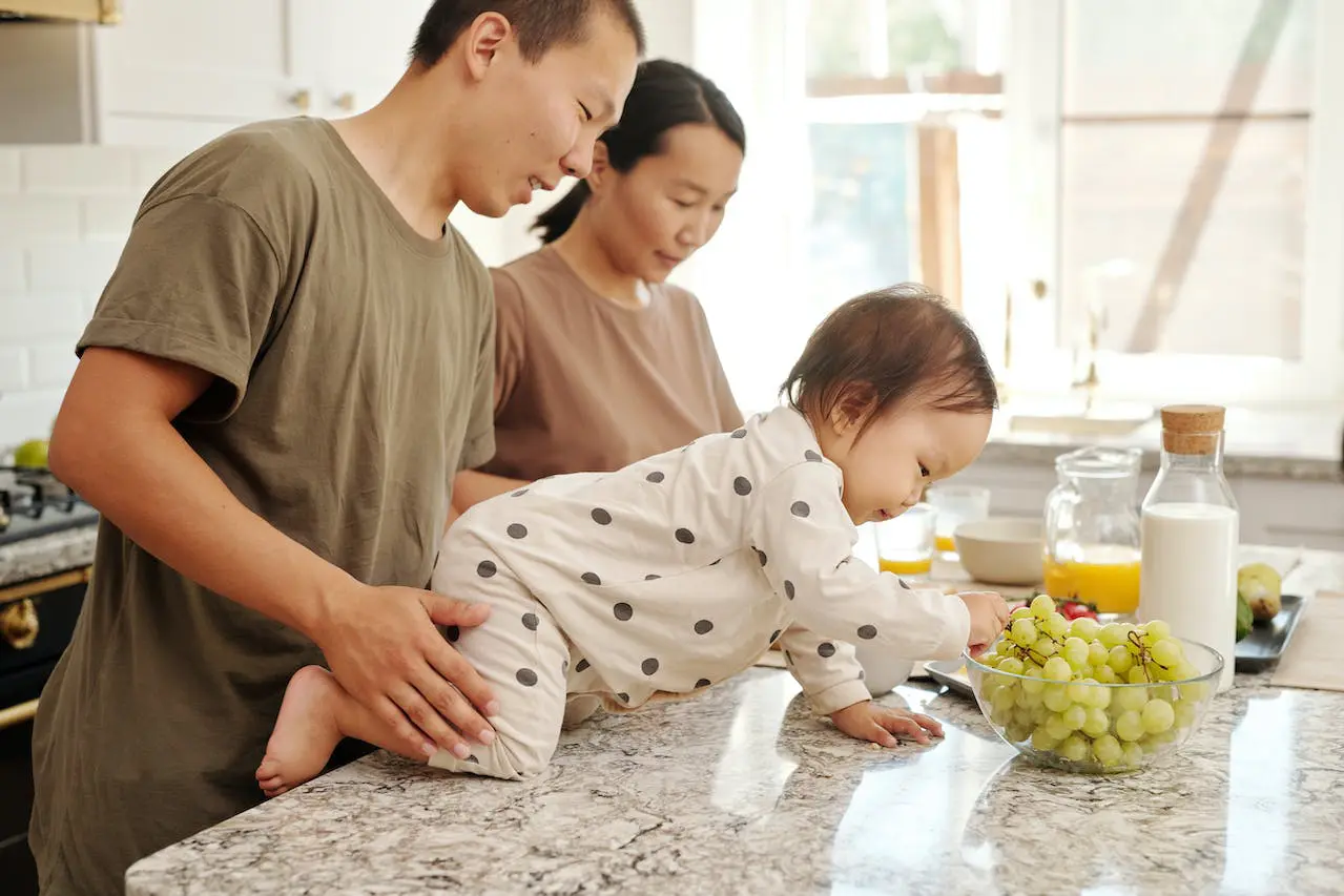 一位父亲抱着一个婴儿，婴儿在柜台上爬向葡萄，母亲在他们旁边做饭，